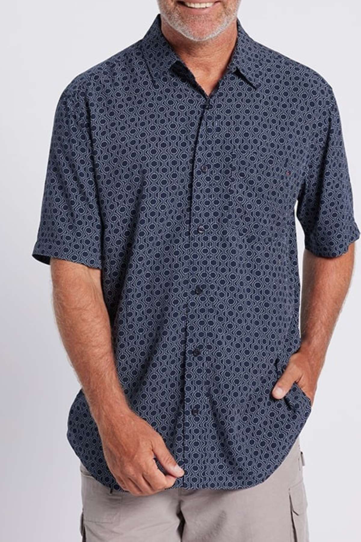 A person with a light beard and mustache is smiling, wearing a short-sleeve Breakaway Jac Bamboo Shirt in dark blue paired with beige cargo shorts. Their right hand rests in their shorts pocket, while the left hand hangs relaxed by their side against a plain white background.