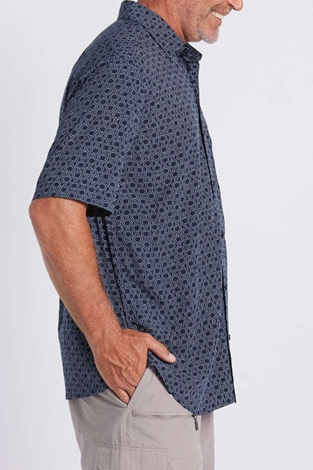 A man is standing sideways with his right hand in the pocket of his light gray shorts. He is wearing a short-sleeved Breakaway Jac Bamboo Shirt in dark blue geometric print and smiling slightly. The background is plain white.