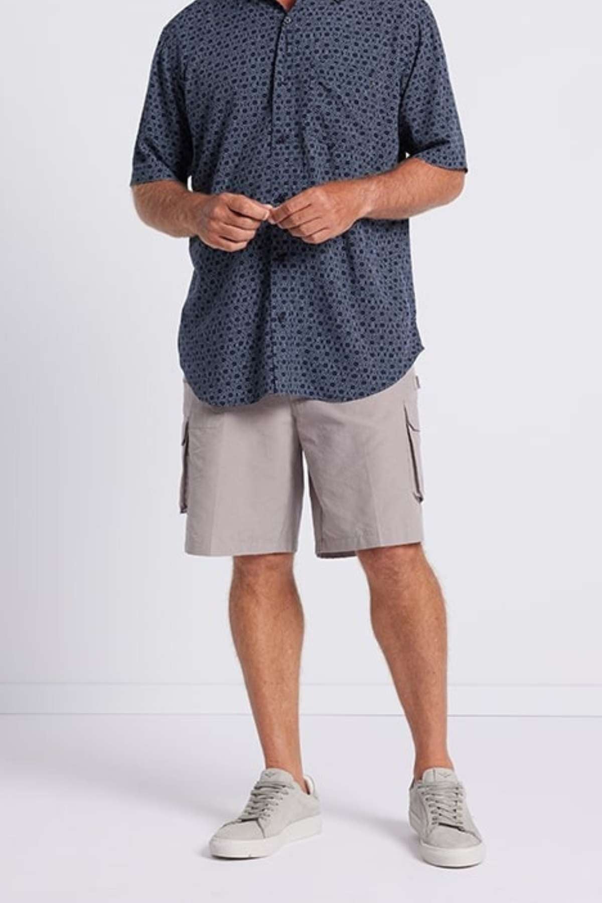 A person wearing a blue geometric print Breakaway Jac Bamboo Shirt, light gray cargo shorts, and gray sneakers is standing against a plain white background. They are holding a pair of glasses with both hands. The upper part of the person's face is out of frame.