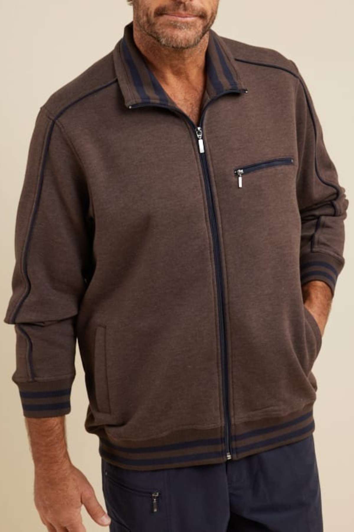 A man models the Breakaway Kyler Snowy Mt Fleece Jacket, a cozy brown zip-up with navy trim, against a beige background. The jacket has a zippered chest pocket and two side pockets. His hands are tucked into the jacket as he wears navy pants, his face partially visible.