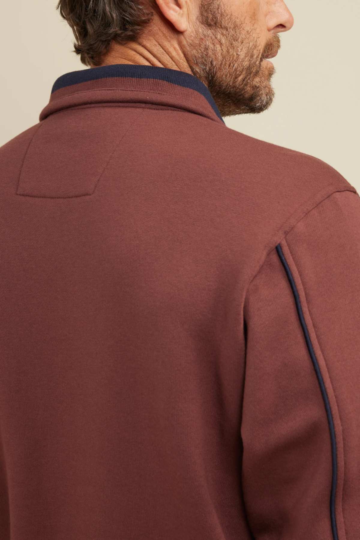 A man with short brown hair and stubble is seen from behind, wearing a maroon Breakaway Kyler Snowy Mt Fleece Half Zip jacket with a dark blue collar. The background is a plain, light beige color. The focus is on the stylish upper back and shoulders of the soft and warm fleece jacket, perfect for winter.