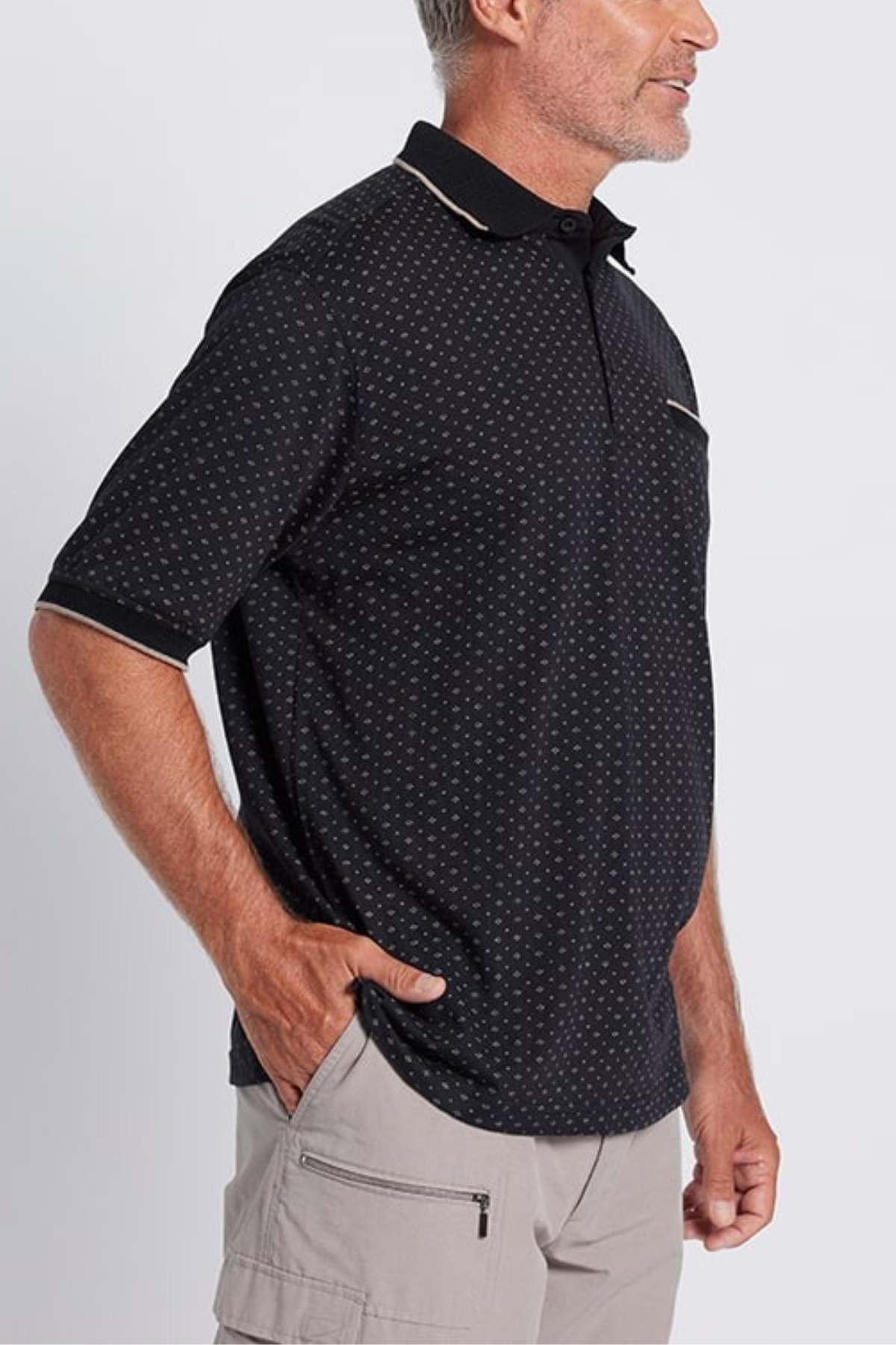 A man with short gray hair stands facing sideways, with one hand in his beige cargo shorts pocket. He is wearing the Breakaway Pete Kinnersly Polo, which features a subtle white geometric print and light beige trim on the collar and sleeves. The background is plain white.