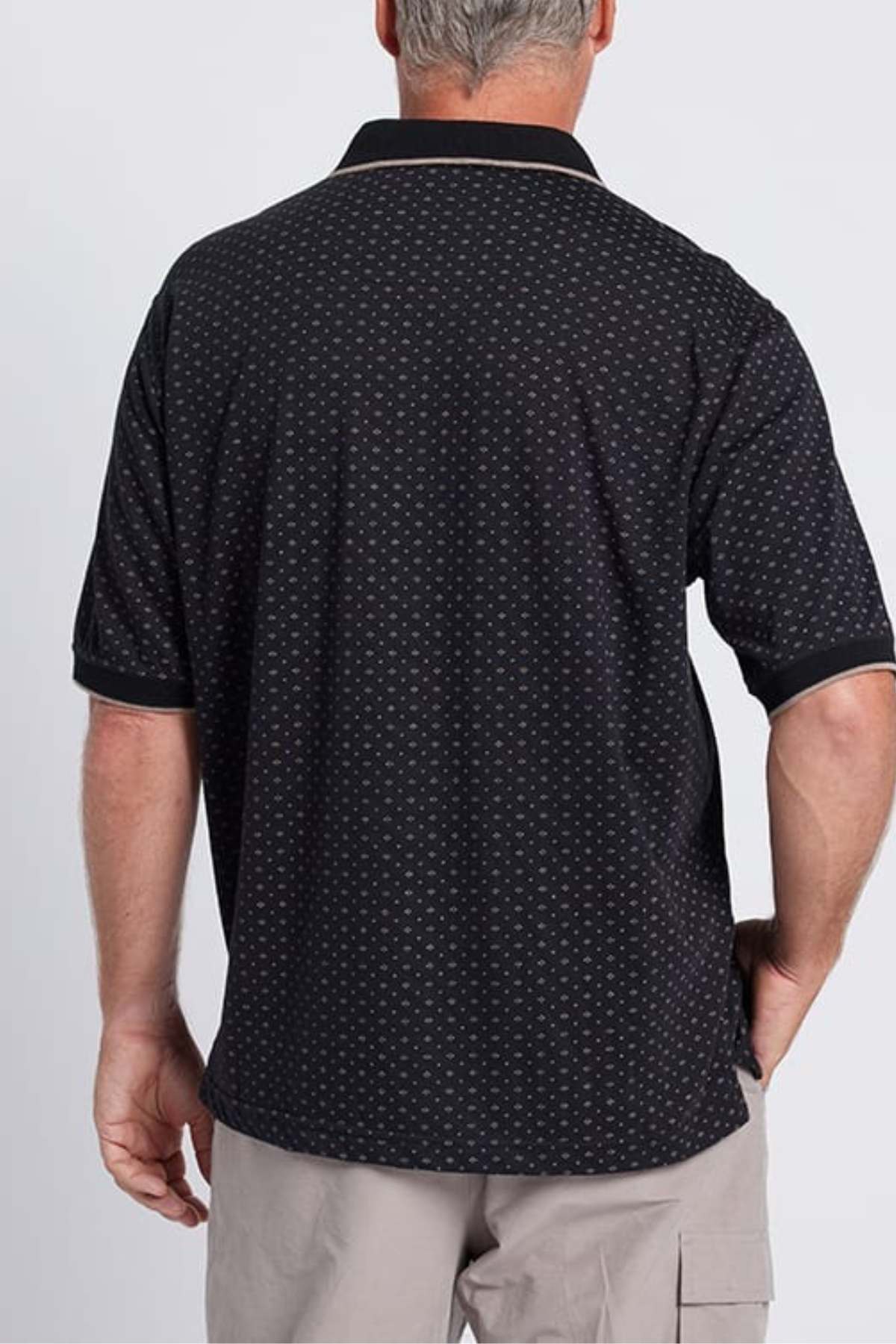 A man with short gray hair is shown from the back, wearing the Breakaway Pete Kinnersly Polo in black, featuring a subtle geometric print paired with khaki shorts. The background is plain and light-colored.