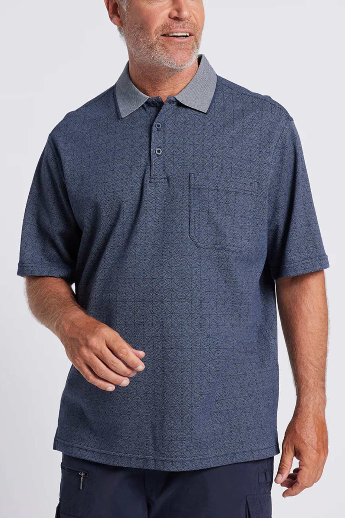 A man with short gray hair and a beard wears the Breakaway Rye Micro Prima Polo in Denim Marle with a birdseye check, light gray collar, and chest pocket. He smiles and looks left against a plain light gray background.