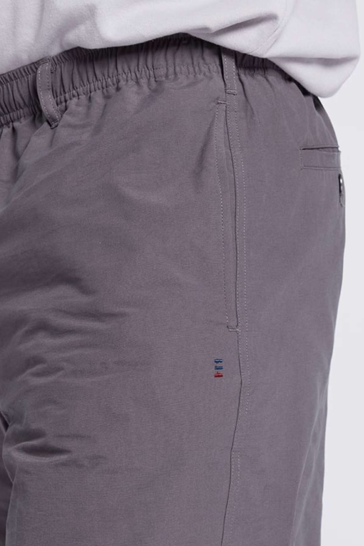 Close-up of a person wearing gray Breakaway Woodbury Short with an elastic and adjustable waistband and a side pocket. Near the pocket seam, there is a small embroidered design featuring three vertical lines in different colors. The edge of a light-colored shirt is partially visible above the waistband.
