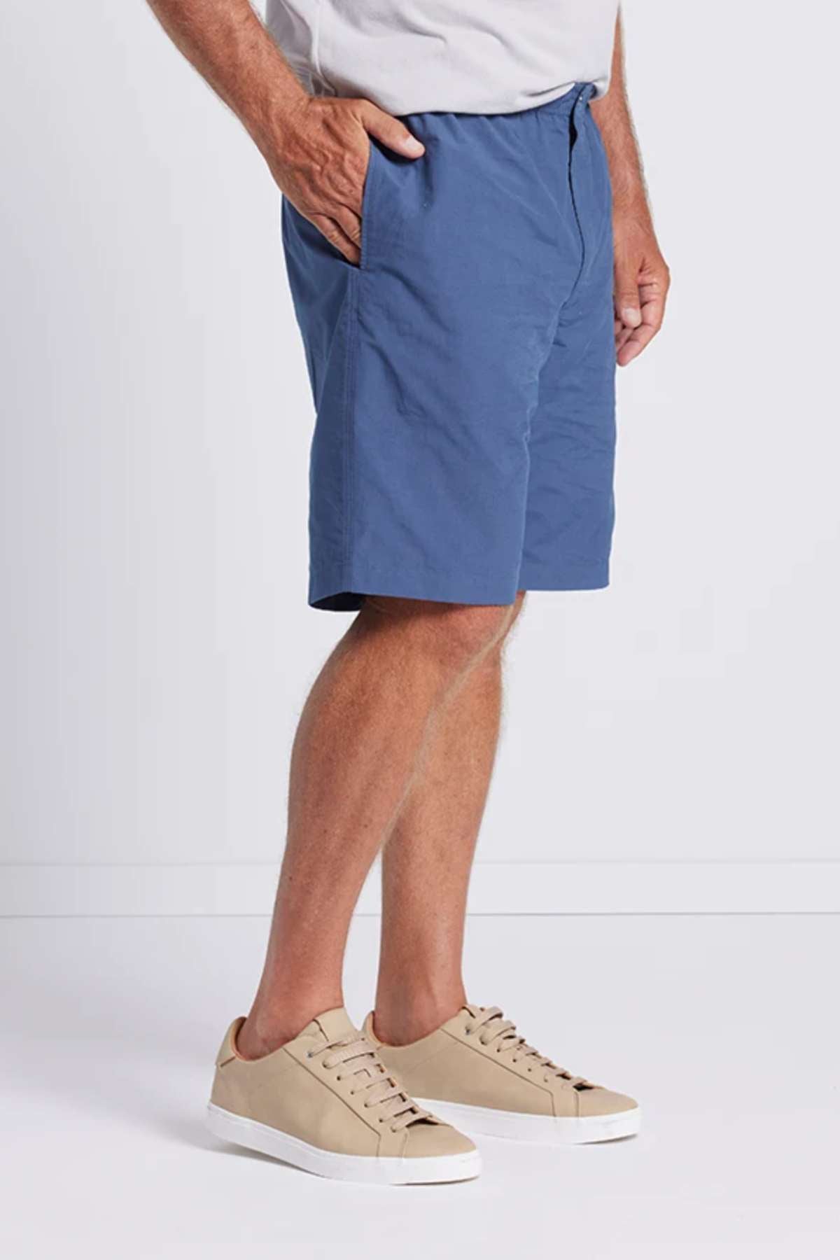 A person is wearing a light gray t-shirt, blue Breakaway Woodbury Shorts with an adjustable waist, and light tan sneakers. They have one hand in their pocket and are standing in front of a plain white background. Only the lower half of the person's body is visible.