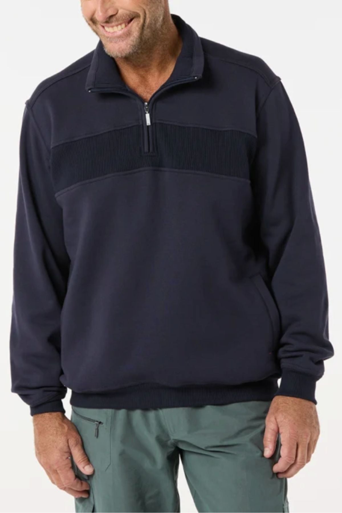 A man wears the Breakaway Zed Snowy Mt Fleece Half Zip— a dark navy pullover with ribbed chest detailing and cozy fleece lining—paired with light green pants, standing against a plain light background and smiling.