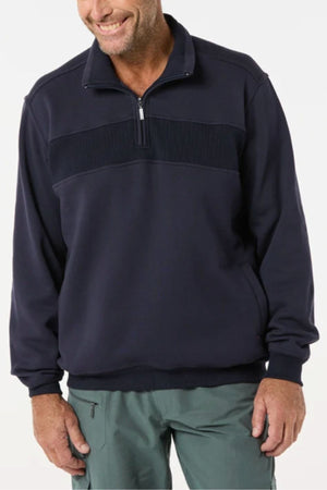 A man wears the Breakaway Zed Snowy Mt Fleece Half Zip— a dark navy pullover with ribbed chest detailing and cozy fleece lining—paired with light green pants, standing against a plain light background and smiling.