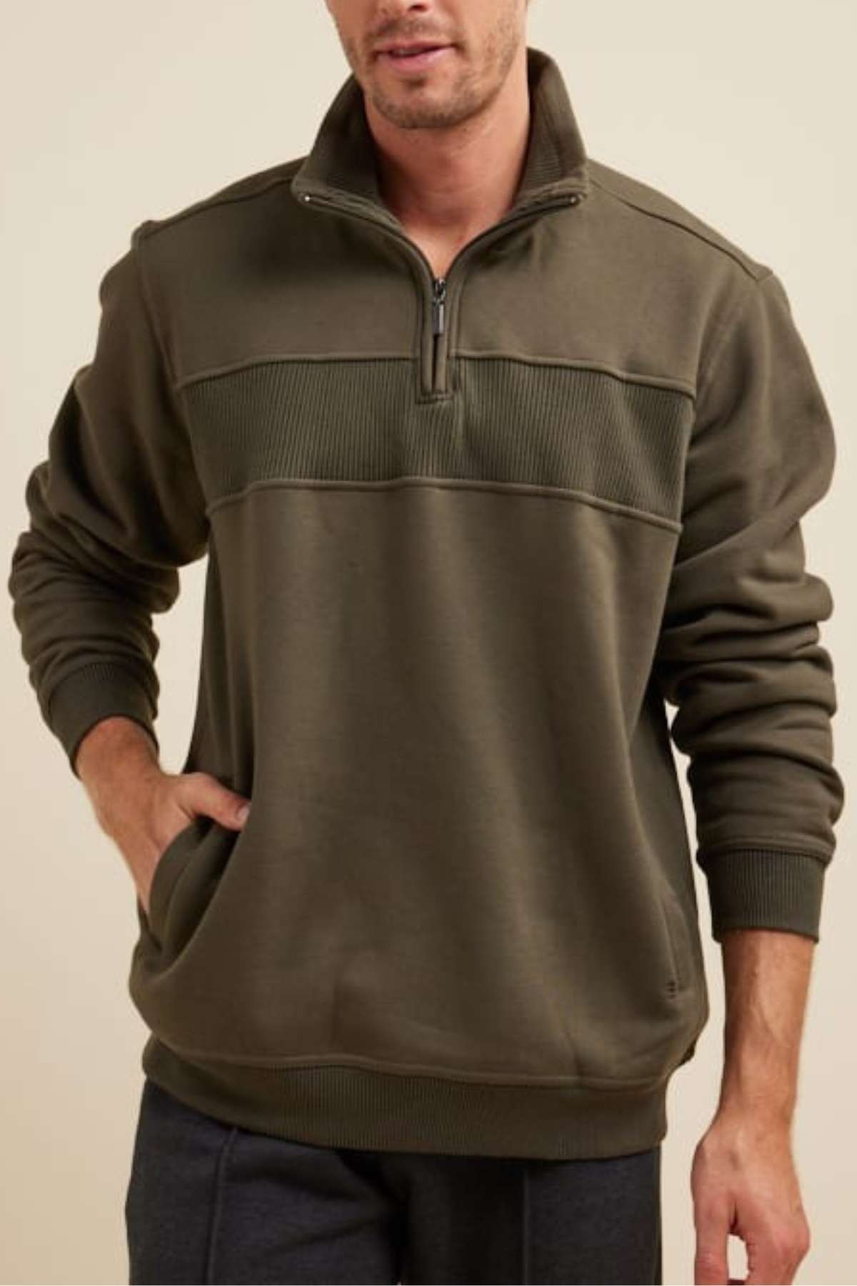 A person with short hair is wearing an olive green Breakaway Zed Snowy Mt Fleece Half Zip pullover with a ribbed texture on the upper chest and shoulders. They have one hand in their pocket, and the background is plain beige.