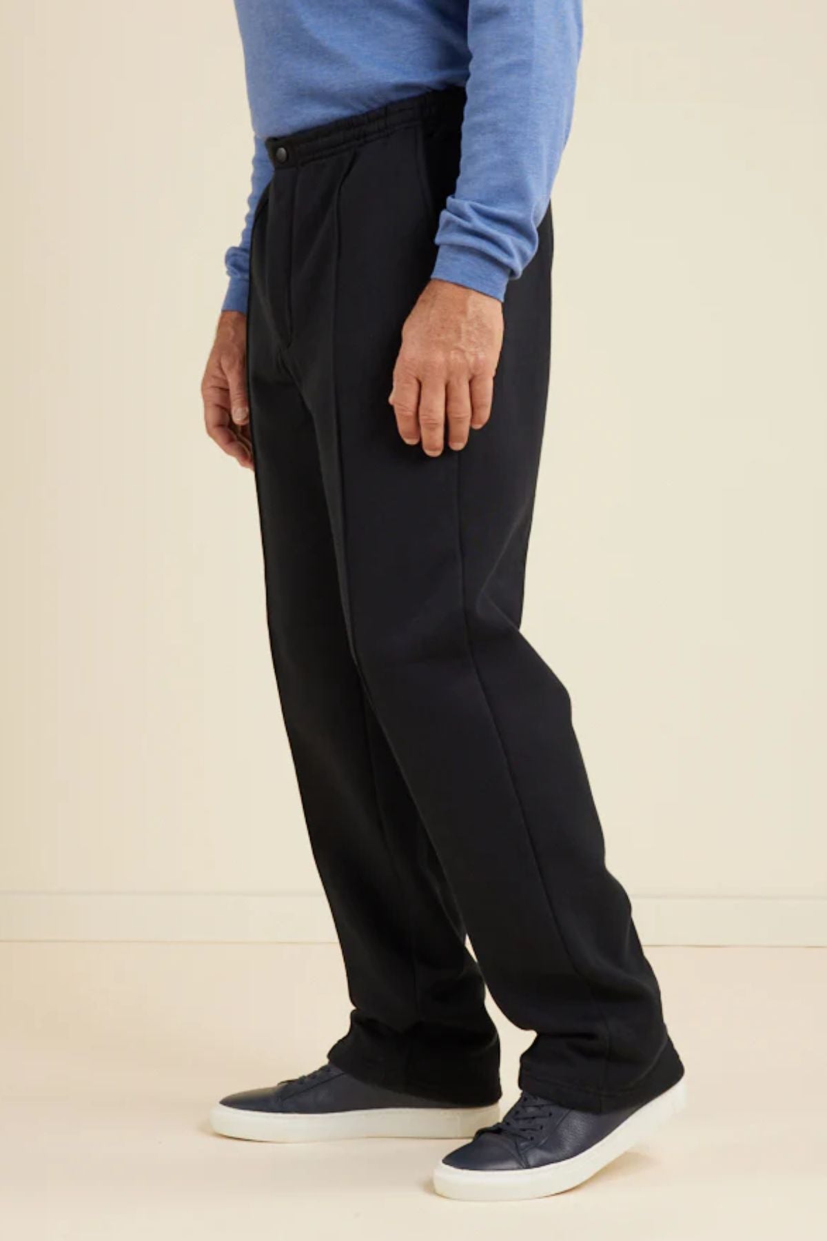 A person wears the Breakaway Pocket Snowy Mt Fleece Pant—black with an elasticated waist—paired with a blue long-sleeve shirt and navy sneakers, standing against a plain light backdrop. The photo is cropped from the shoulders down.