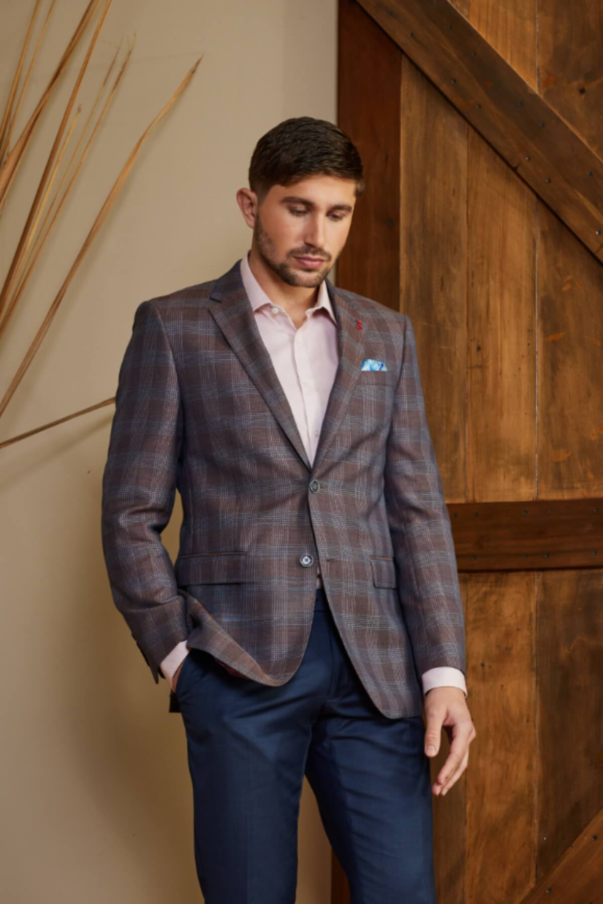 A man poses indoors in a Bruton Abram Linen Blazer, complemented by a light pink shirt and dark trousers. With short hair, he rests one hand in his pocket while standing against a wooden door and beige wall decorated with sticks.