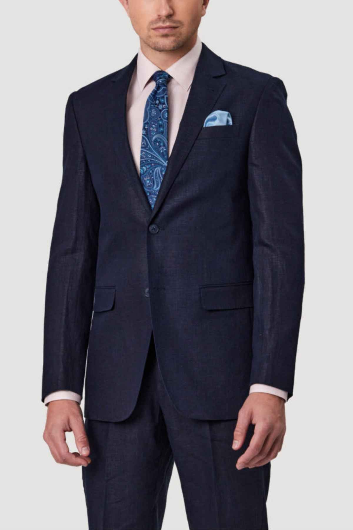 A man dressed for summer occasions wears a dark suit paired with a light pink shirt and a blue paisley tie. The ensemble is complemented by the Bruton Abram Pure Linen Blazer, while a light blue pocket square adds a touch of flair. The plain, light-colored background provides a sophisticated setting.