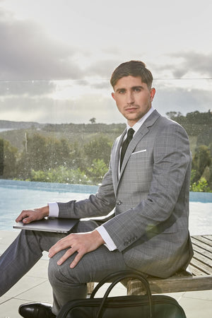 A man in a Savile Row Abram Blazer sits on a bench near a pool, holding a laptop. He has short brown hair and appears focused. The background features greenery and an overcast sky. A black bag rests beside him.