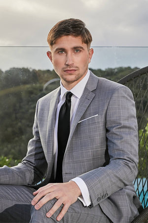 A man with short brown hair and a trimmed beard is sitting on a modern outdoor chair. He is wearing the Savile Row Abram Blazer over a white dress shirt and black tie. The background features a scenic, hilly landscape under a cloudy sky.