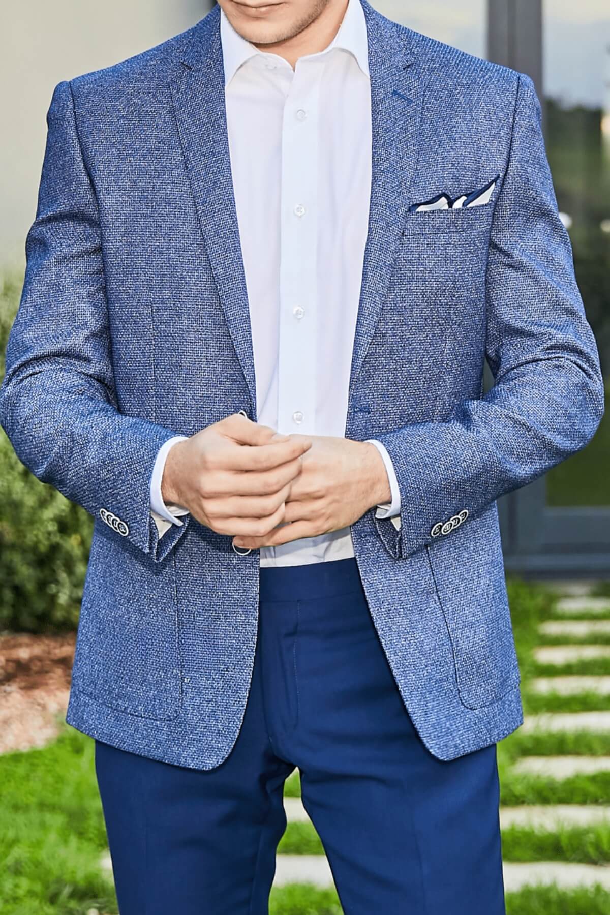 Someone stands outside in a light blue Bruton Adin Blazer, paired with a white dress shirt and dark blue pants. Hands are clasped; a pocket square adds detail. The geometric texture of the blazer complements the greenery and building behind them.