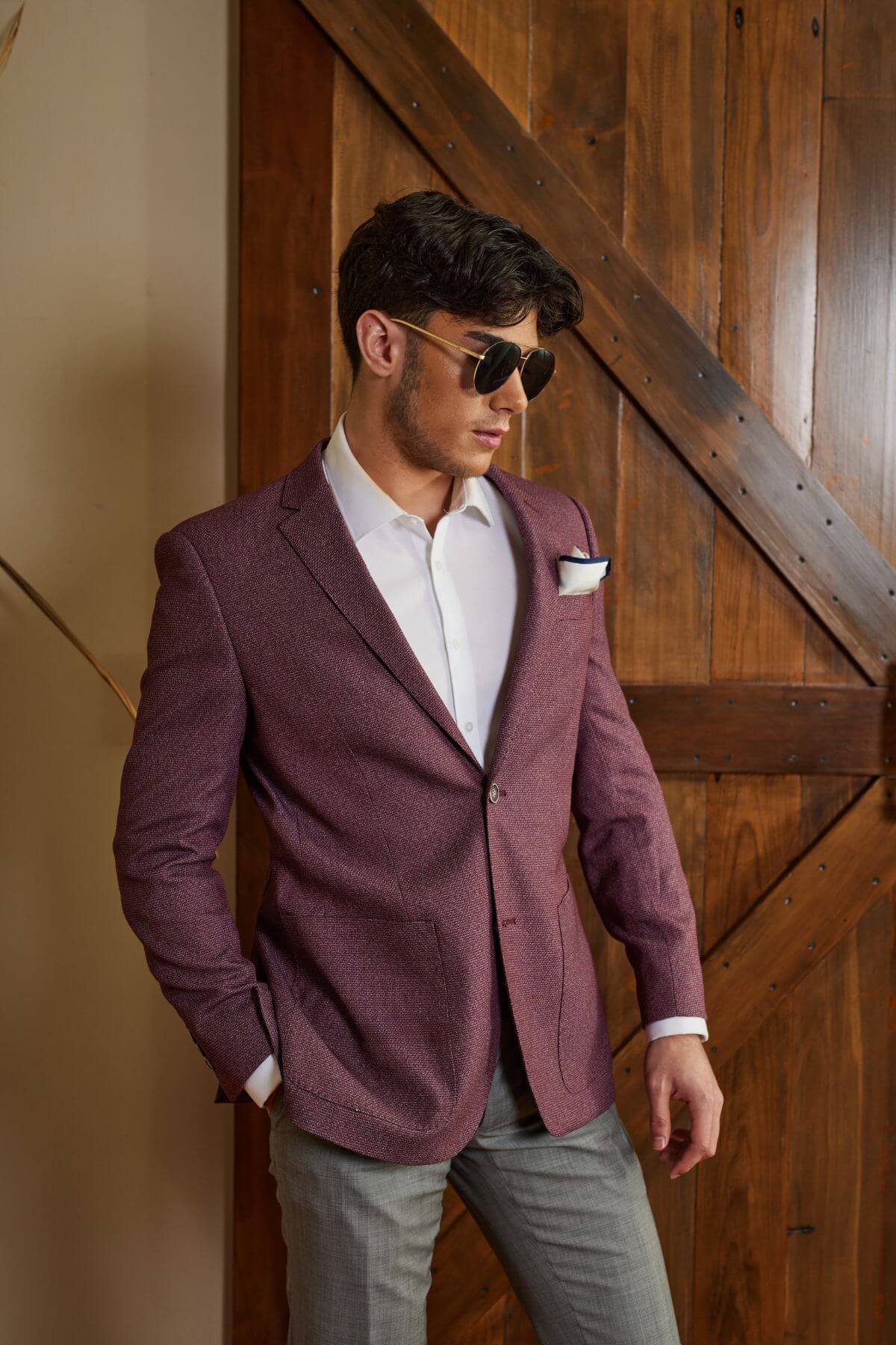 A man wearing sunglasses, a white shirt, and the Bruton Agar Blazer, complete with a pocket square, stands in front of a wooden door accented with diagonal metal braces. With the blazer's tailored look and modern fit, he appears relaxed, one hand in his pocket and the other resting on the door.
