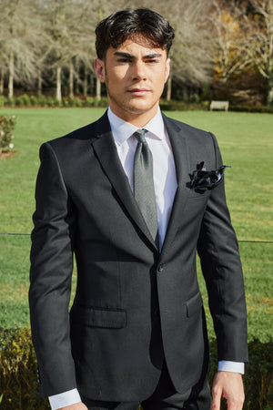 A young man stands outdoors wearing a Bruton David Suit Jacket, a white dress shirt, and a patterned tie. His dark hair is neatly styled, and in his breast pocket is a black pocket square. The background features a grassy area, trees, and a bench.