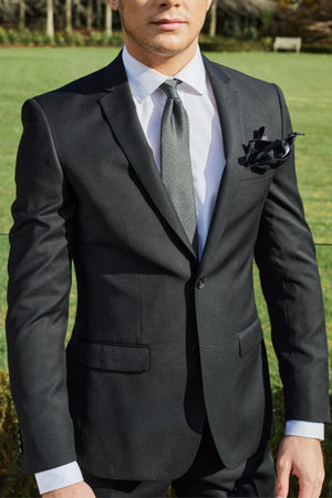 A person wearing a Bruton David Suit Jacket in black, paired with a white dress shirt and grey tie, stands outdoors. A black pocket square accents the breast pocket of the slim-fit suit jacket. The background showcases a lush green lawn and trees, while the person's head is slightly cropped out of the image.