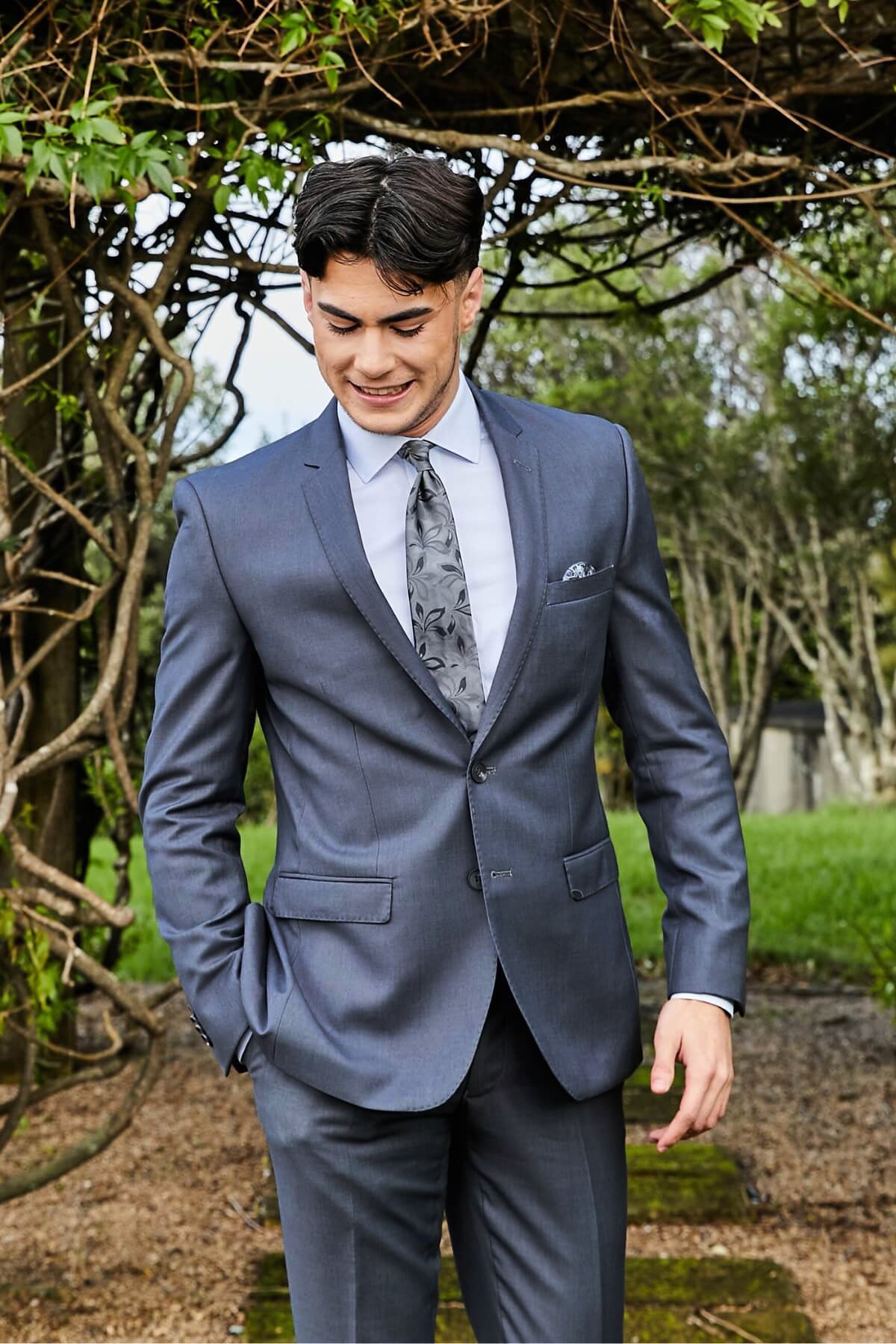 A young man wears the Bruton David Suit Jacket and a patterned tie, smiling outdoors with one hand in his pocket against a backdrop of leafy greenery.