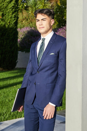 Wearing the Bruton David Suit Jacket, a person stands outside with a laptop, leaning against a light pillar. The structured shoulders of the blue, slim-fit jacket are enhanced by a lush green and purple-flowered bush in the background.