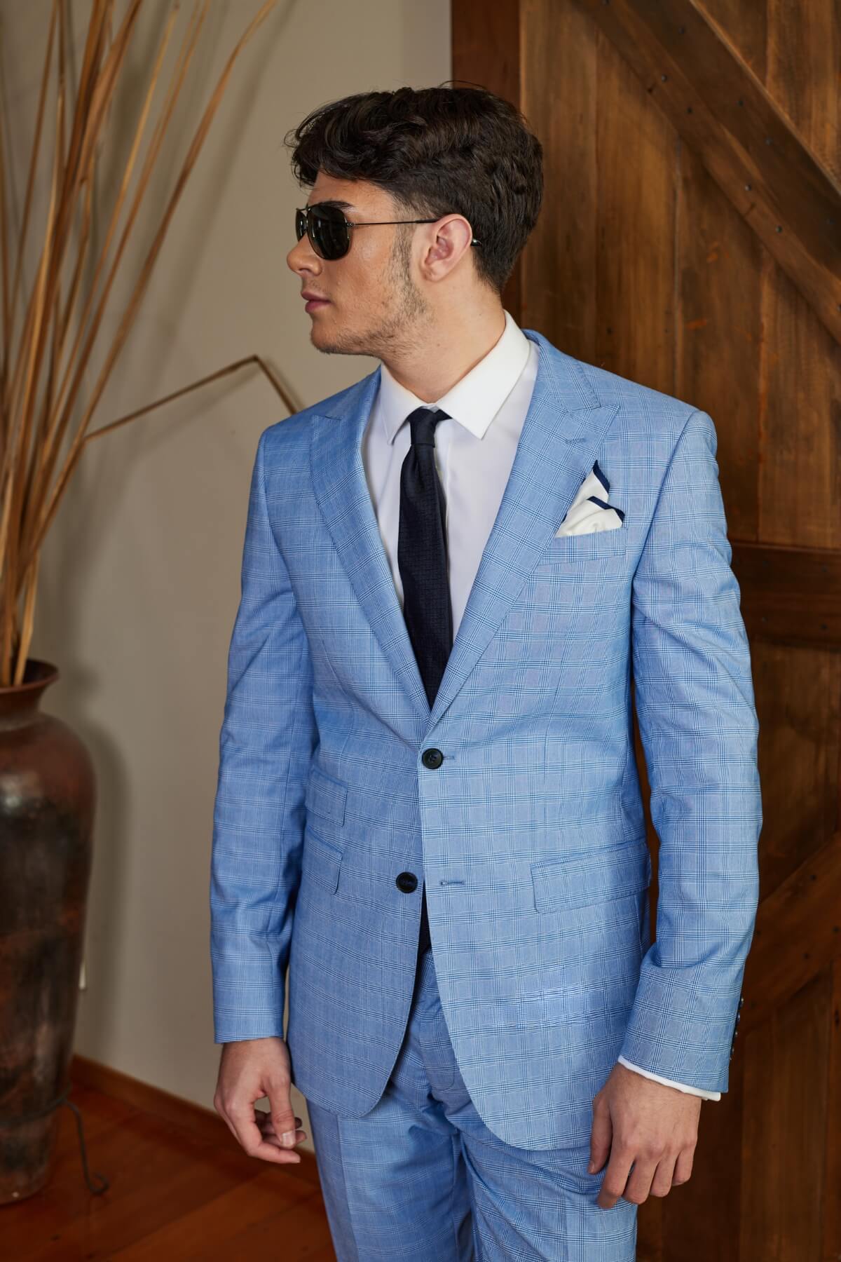 A man wearing a light blue Bruton Jed suit jacket with peak lapel, white shirt, dark tie, sunglasses, and a pocket square stands indoors. A wooden door and tall decorative plant are visible in the background.