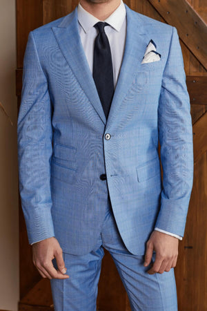 A person dons a slim-fit Bruton Jed Suit Jacket in light blue, featuring a peak lapel, paired with a white dress shirt and dark tie, in front of a wooden backdrop. A pocket square is neatly tucked into the breast pocket, but the person's face is not visible.