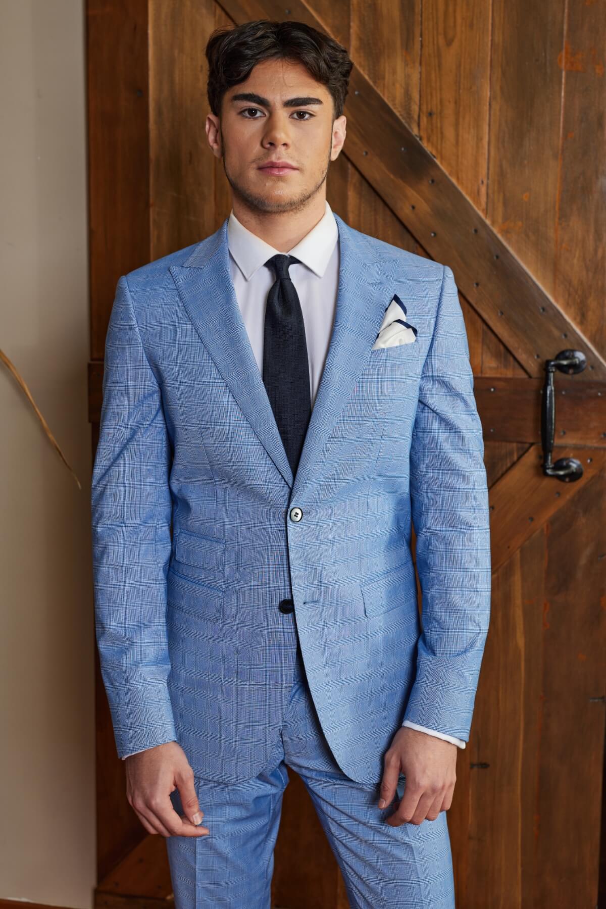 A man stands neutrally before a wooden door, wearing a light blue Bruton Jed Suit Jacket featuring peak lapels. He's paired it with a white dress shirt and black tie, has one hand slightly raised, and a handkerchief tucked into the pocket of his slim-fit jacket.
