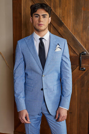 A man stands neutrally before a wooden door, wearing a light blue Bruton Jed Suit Jacket featuring peak lapels. He's paired it with a white dress shirt and black tie, has one hand slightly raised, and a handkerchief tucked into the pocket of his slim-fit jacket.