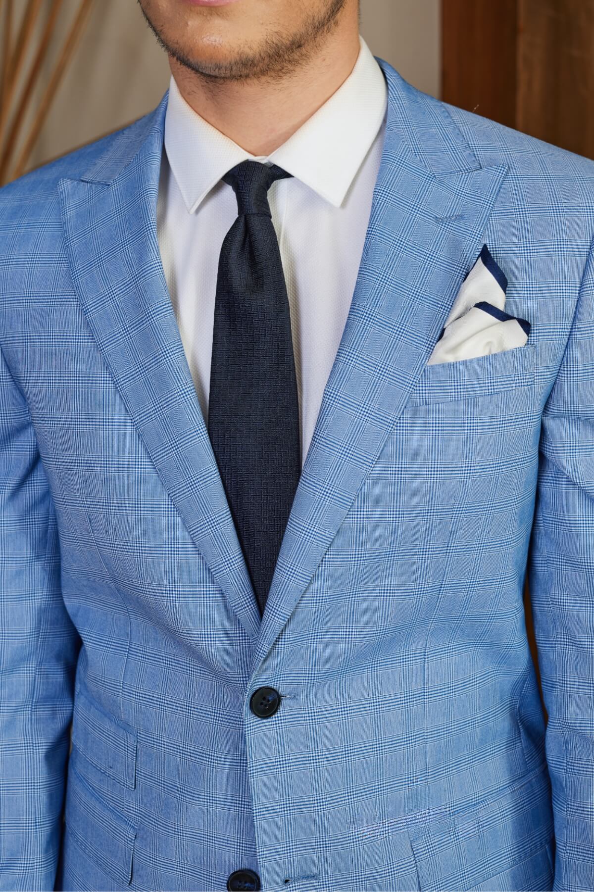 A person in a slim fit light blue checkered suit with a white shirt and dark tie, wearing the Bruton Jed Suit Jacket featuring a peak lapel and a neatly placed white pocket square. The background is softly blurred.