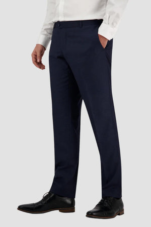 Dressed in Bruton Jesse Trousers with their left hand in a pocket, a person exudes elegance through the fine check print of the slim-fit pants paired with a white shirt and black leather shoes, set against a plain light gray background.