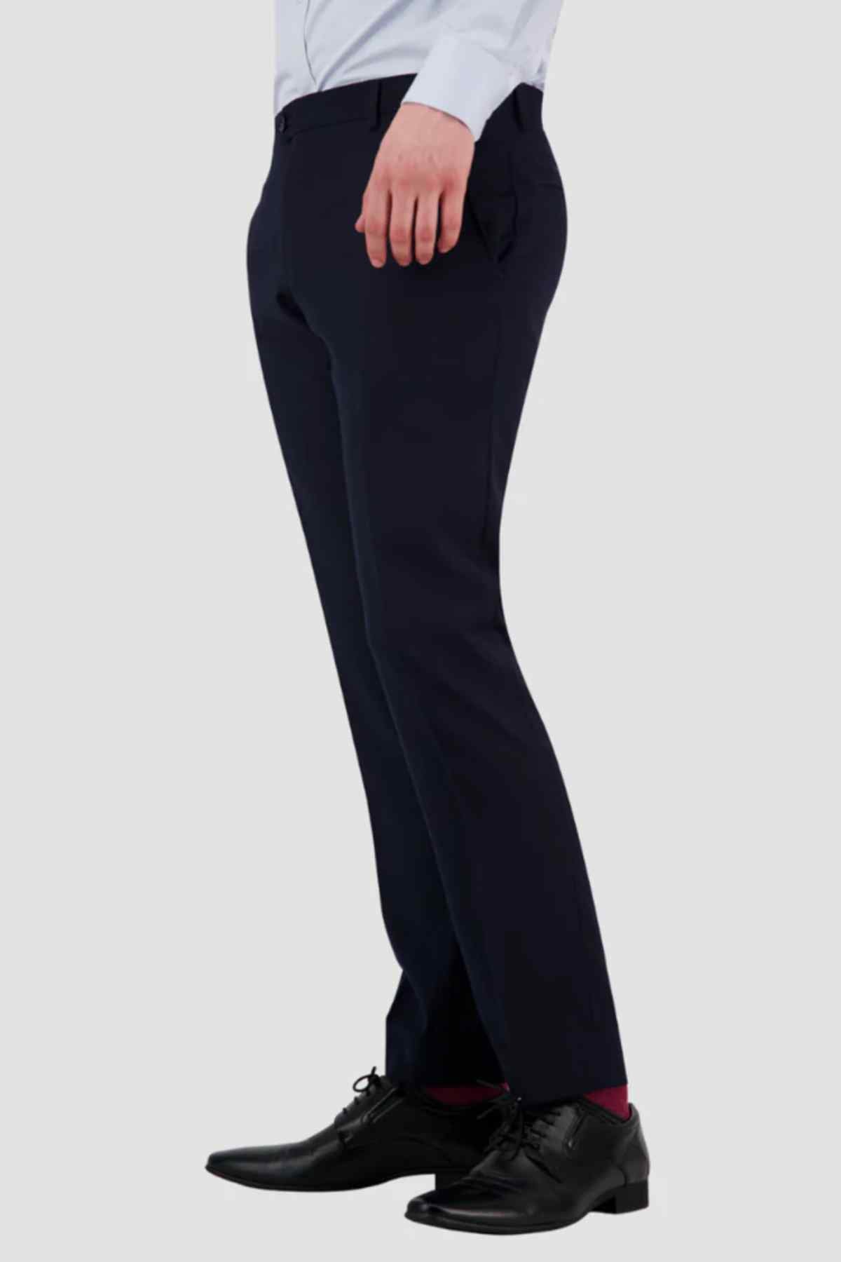 A person wearing Bruton Jesse Trouser with button back pockets, a light blue shirt, black dress shoes, and red socks stands against a plain light gray background. Only the lower half of the body is visible.