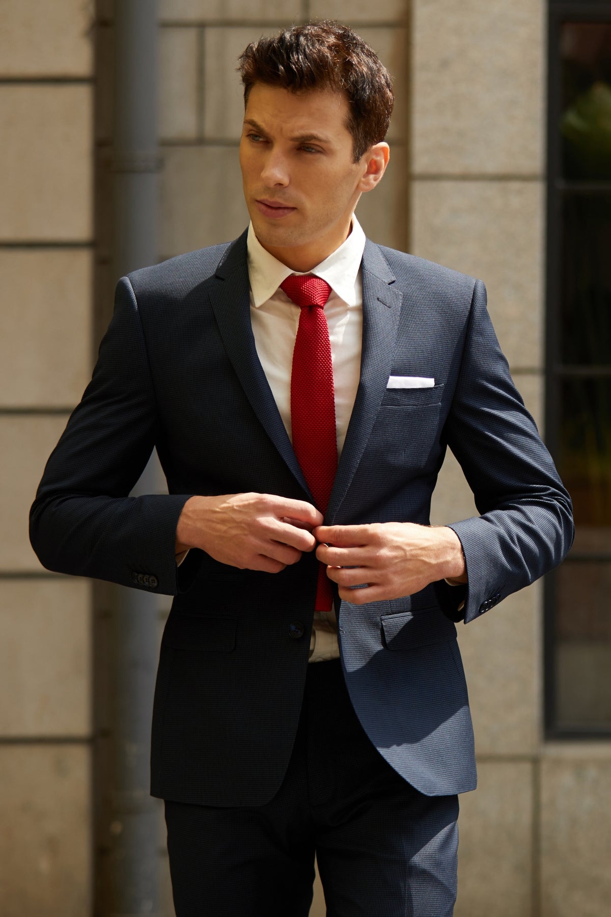 A man wearing a slim fit Bruton Jose Suit Jacket, paired with a red tie and white pocket square, fastens his jacket outdoors in front of a light stone building, highlighting a sharp professional style.