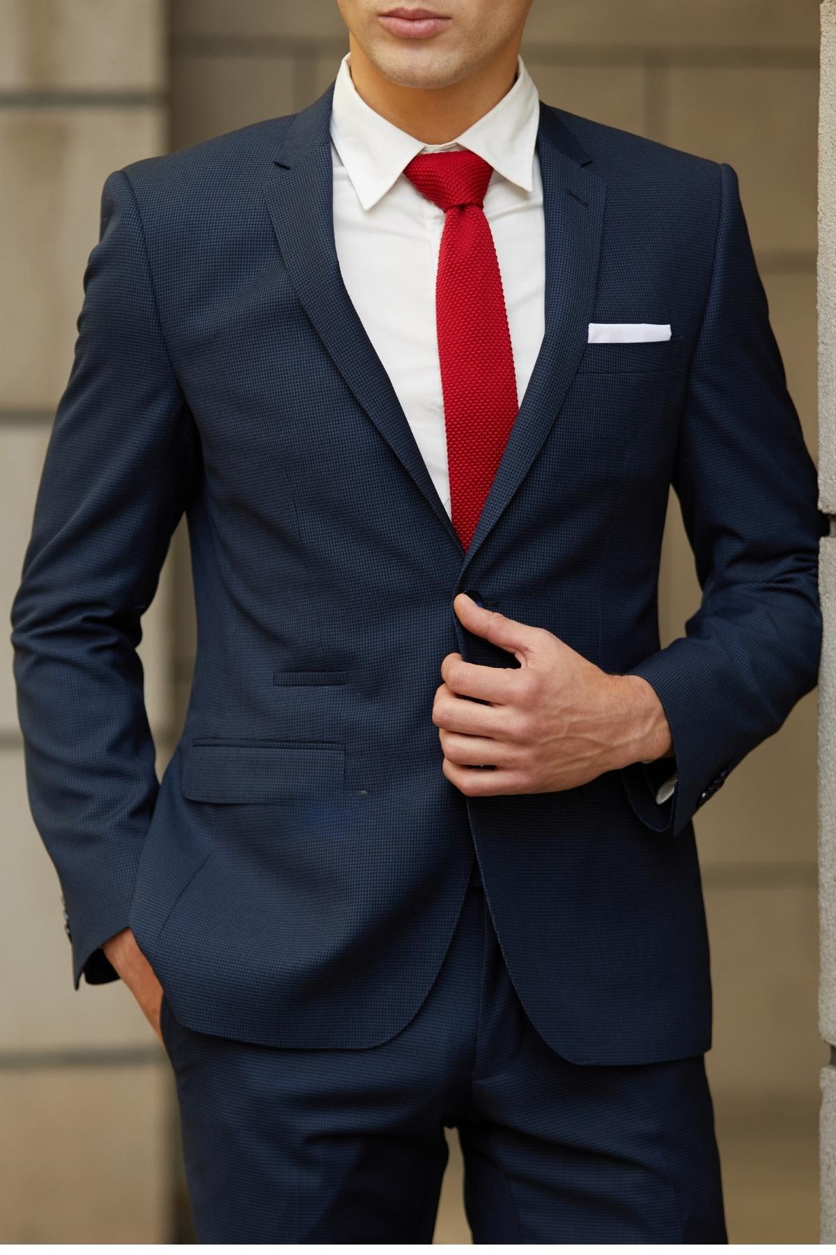 A man in a Bruton Jose Suit Jacket, paired with a white shirt, red tie, and white pocket square, stands with one hand in his pocket and the other adjusting his jacket. His face is partly visible against a blurred background.