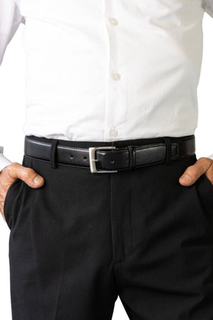 A person in a white dress shirt tucked into black pants wears the Buckle 5115 Rogue Deluxe Leather Belt with a brushed nickel buckle, hands in pockets.