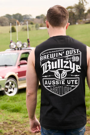 Wearing the Bullzye Brewin Dust Muscle Tank with its bold “Bullzye Aussie Ute Nation” graphic, a man stands in a grassy field beside a ute equipped with roof spotlights. This muscle tank is an essential for men’s casual style.