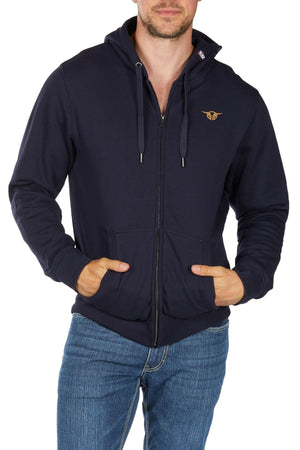 A man wearing the Bullzye Hardy Zip Up Hoodie in navy with a small chest logo and blue jeans stands with his hands in the hoodie pockets against a plain white background.
