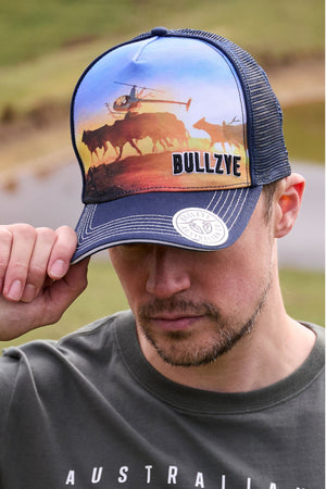 A man touches the brim of a Bullzye Outback High Profile Trucker Cap, which features a colorful print of cattle, a helicopter, and "BULLZYE," while wearing a dark t-shirt with "AUSTRALIA" partially visible.