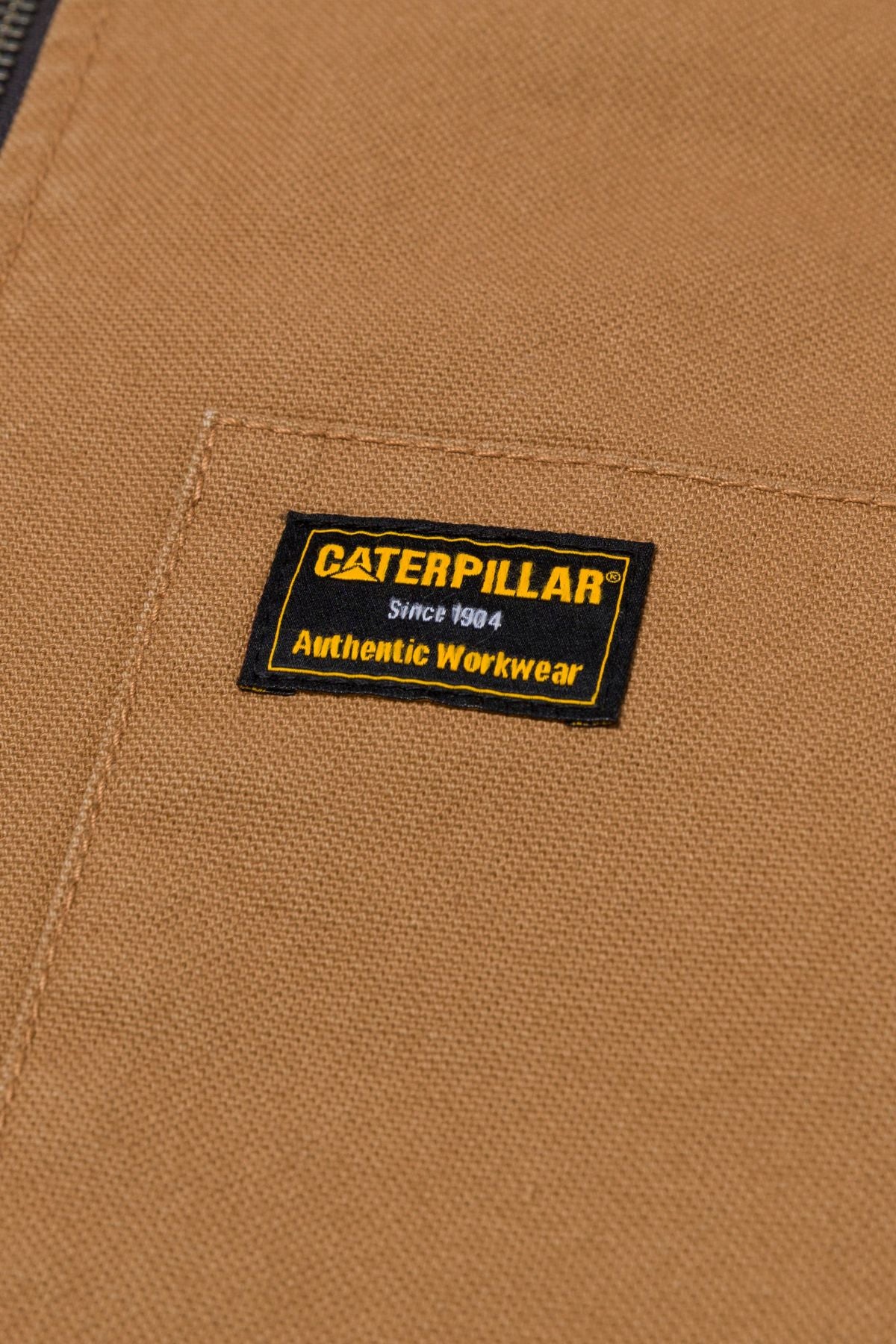 Close-up of the CAT Canvas Utility Sherpa Lined Vest in brown, featuring a stitched black and yellow label reading "Caterpillar Since 1904 Authentic Workwear.