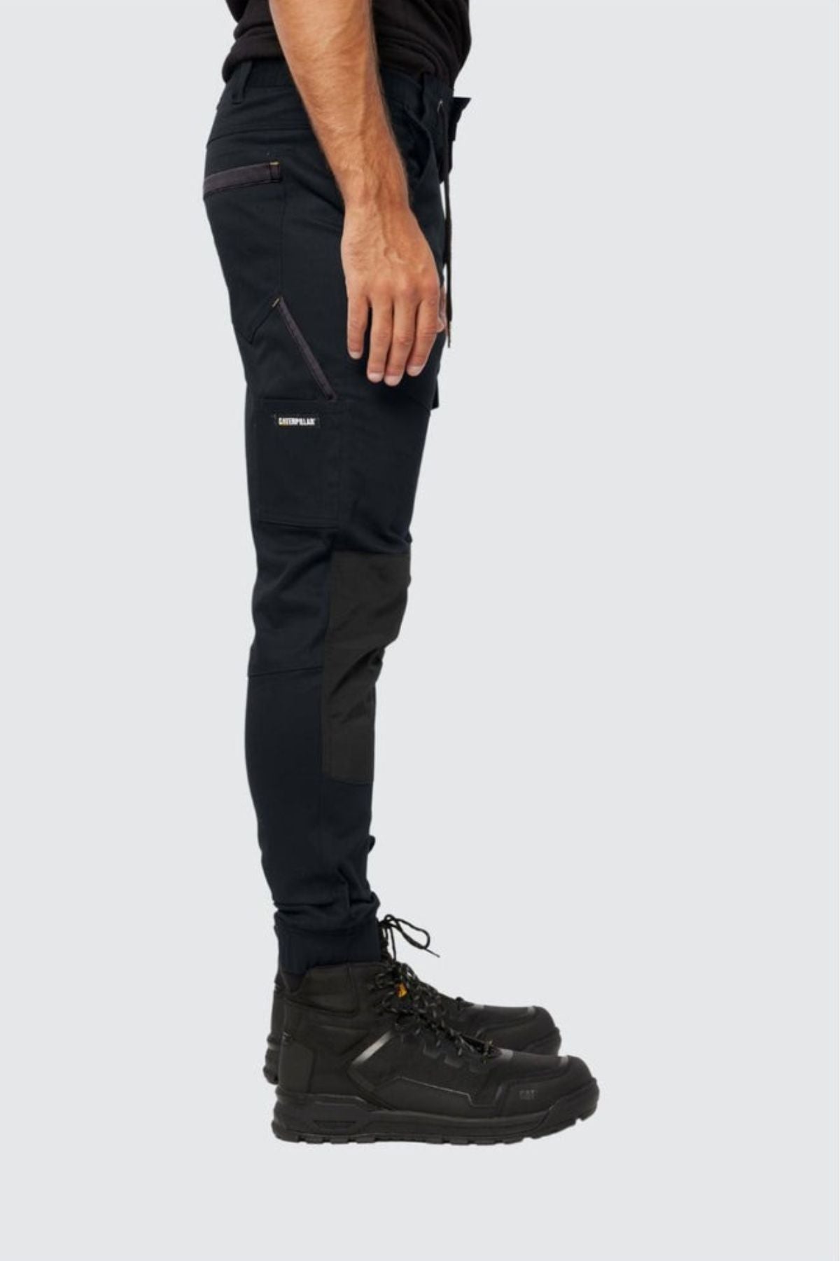 A person stands sideways, wearing CAT Cuffed Dynamic Pant with zipper pockets and black work boots. Only the lower half of the body is visible against a plain light background.