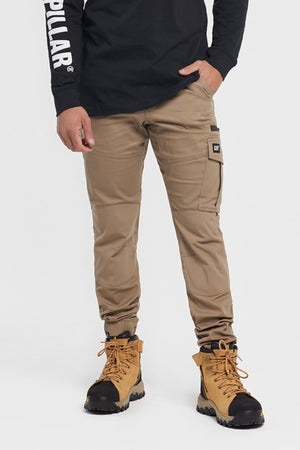 Wearing CAT Cuffed Dynamic Pant in tan and tan hiking boots, a person stands against a plain white background. Only the lower half, including black long-sleeve shirt with white sleeve text, is visible.