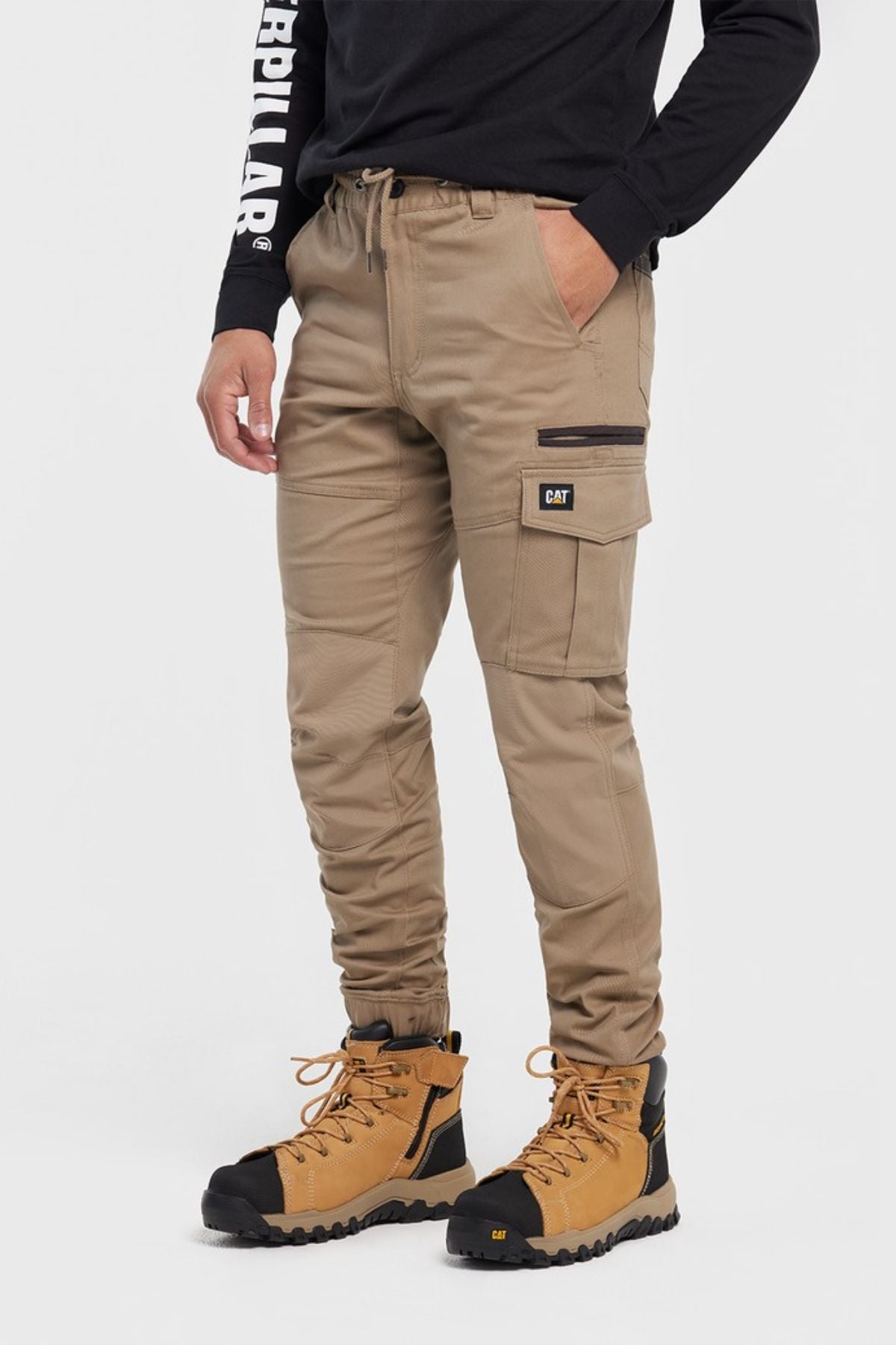A person in CAT Cuffed Dynamic Pant cargo joggers, a black long-sleeve shirt with "Caterpillar" on the sleeve, and tan work boots stands against a plain white background.