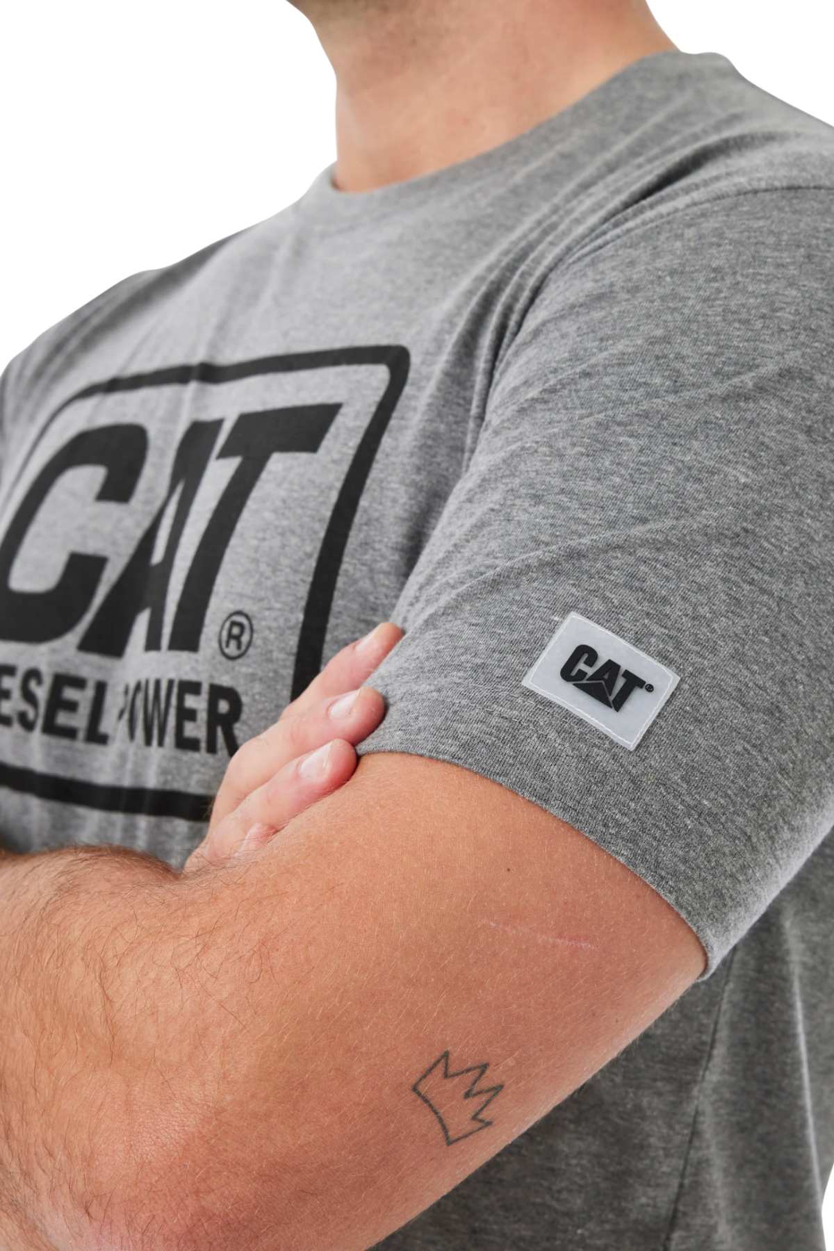 A person with crossed arms and a small crown tattoo on their forearm wears a gray CAT Diesel Power Tee, which features a rib collar and a fabric brand patch on the sleeve.