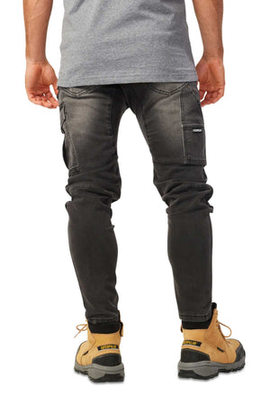 A person is shown from the back wearing dark gray CAT Dynamic Denim Pants with multiple pockets and stitching details, paired with tan work boots and a gray shirt.