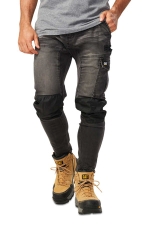 A person wearing stylish CAT Dynamic Denim Pants, faded black with black knee pads, and comfortable tan work boots with black soles stands against a white background. Their gray shirt is partially visible as their right hand rests casually in the pants pocket.