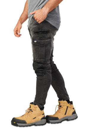 A person is wearing a gray shirt, stylish black CAT Dynamic Denim Pants with multiple pockets, and tan work boots featuring black soles and lace-up details. They're standing against a white background.