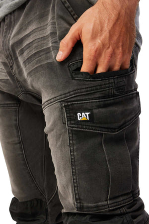 Close-up of a person in CAT Dynamic Denim Pant, showcasing stylish, dark, faded jeans with distinctive seams and stitching. A grey shirt complements the look as they place a hand in the side pocket, highlighting the "CAT" patch that adds to the rugged appeal and comfort.