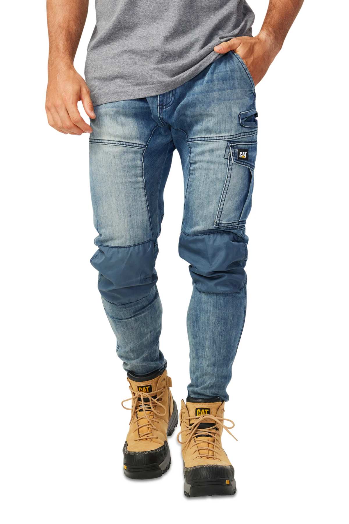 A person in CAT Dynamic Denim Pant, featuring reinforced knees and a cargo pocket, pairs them with tan work boots and a gray t-shirt, standing casually with one hand in their pocket.