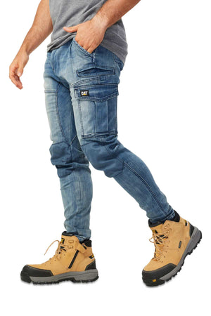 A man, seen from the waist down, stands with one leg forward wearing a gray t-shirt, tan CAT work boots, and blue CAT Dynamic Denim Pant with reinforced knees and multiple cargo pockets.
