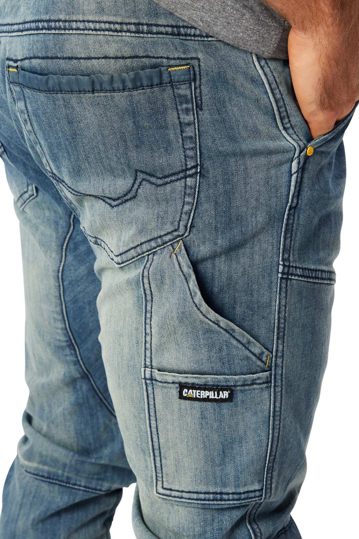 A close-up of a person wearing CAT Dynamic Denim Pant with cargo pockets, visible stitching, and multiple pockets; their right hand is tucked in and a Caterpillar label appears on the side pocket.