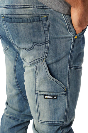A close-up of a person wearing CAT Dynamic Denim Pant with cargo pockets, visible stitching, and multiple pockets; their right hand is tucked in and a Caterpillar label appears on the side pocket.